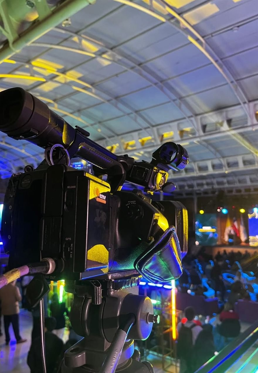 Cámara sobre trípode registrando un evento con público y escenario iluminado.