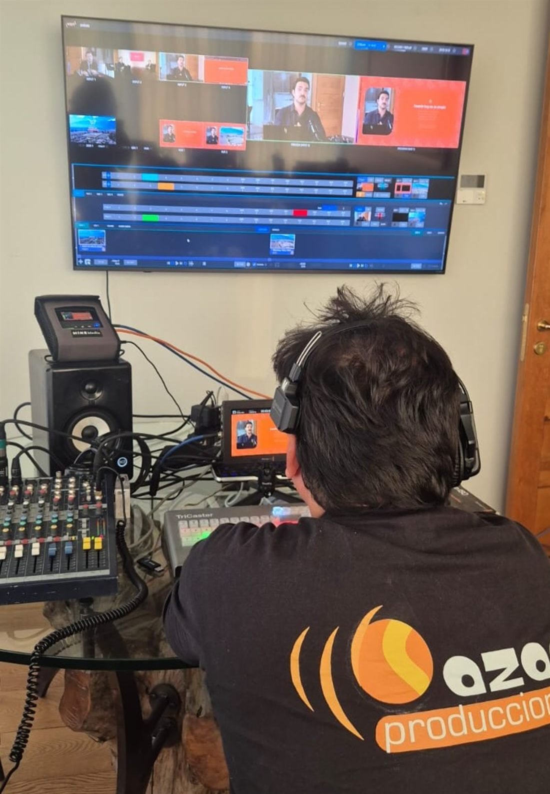 Operador con polera de Azar Producciones frente a una consola y un switcher, monitoreando una transmisión en vivo.