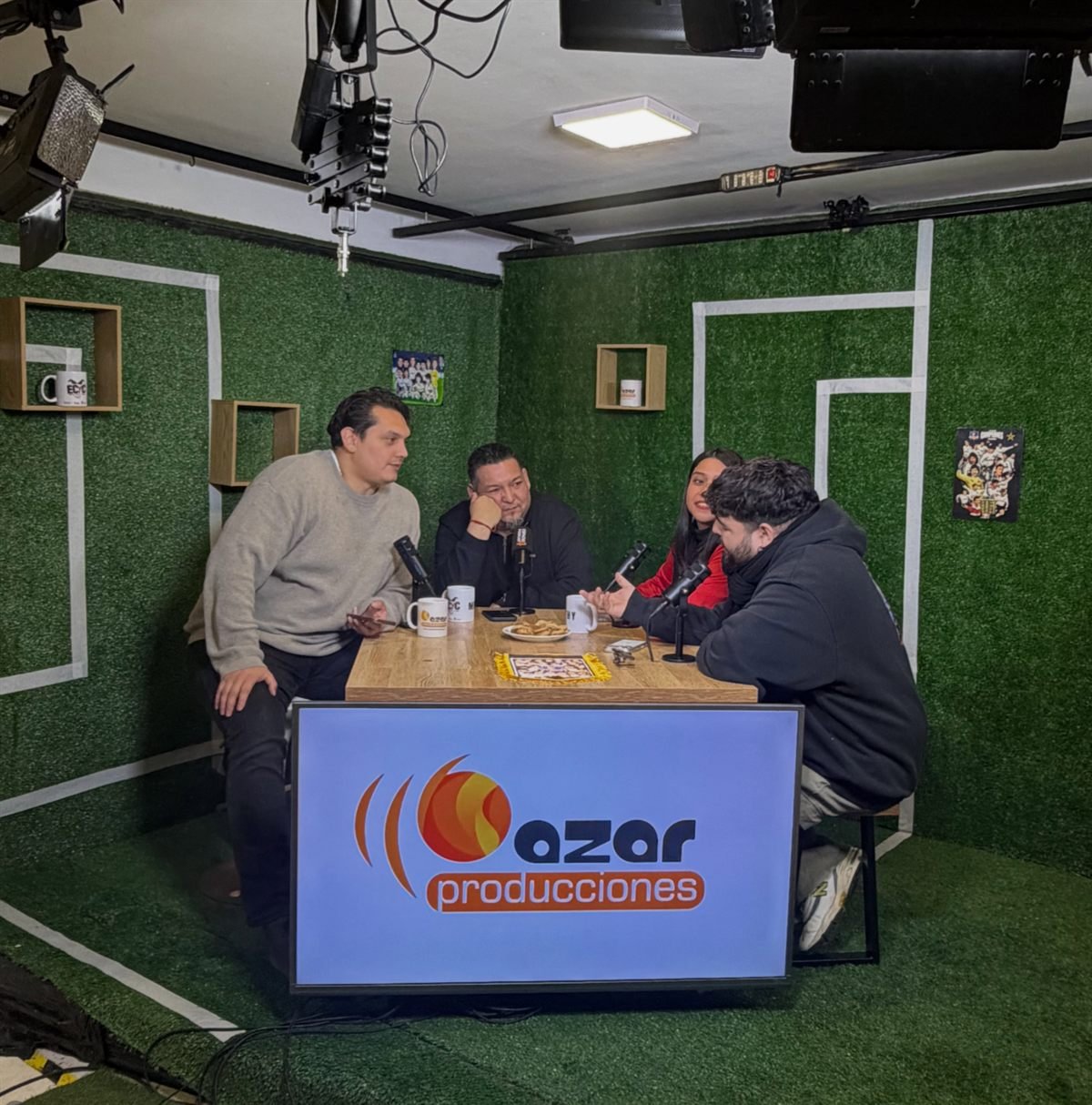 Estudio de podcast con cuatro participantes, micrófonos e iluminación, y pantalla con el logo de Azar Producciones.